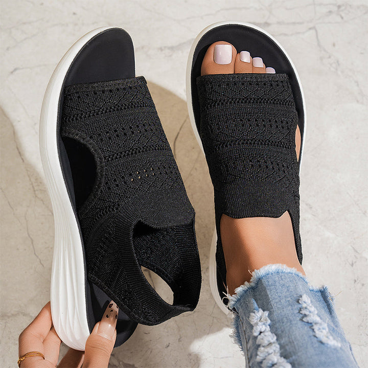Black knit slip-on shoes with white soles worn by a person on a light gray background